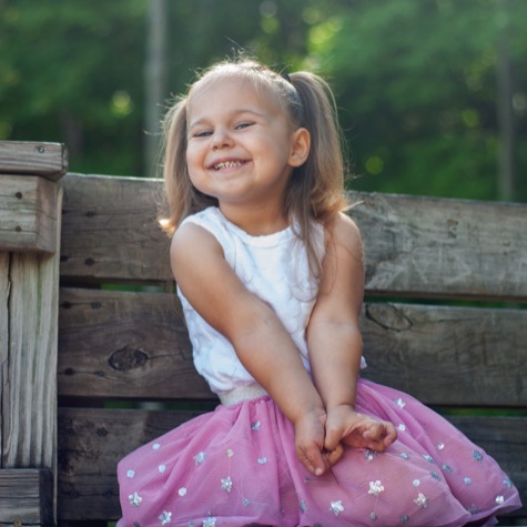 Professional outdoor portrait of a smiling young girl with pigtails wearing a white top and pink skirt while seated on a rustic wooden bench