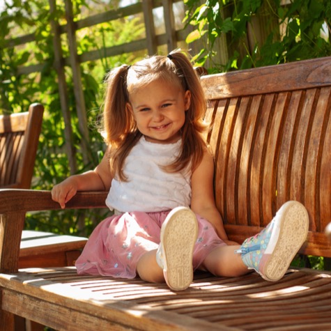 Professional outdoor portrait of a smiling young girl with pigtails wearing a white top and pink skirt while sitting playfully on a sunlit wooden garden bench