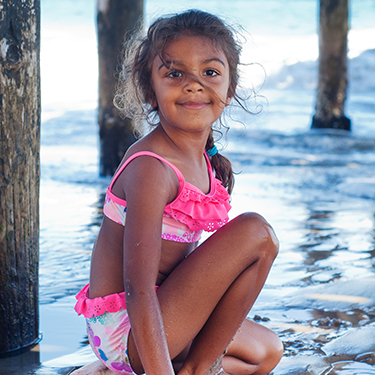 Young girl in a colorful swimsuit smiling and sitting on the beach beneath a rustic wooden pier during an outdoor professional child portrait photography session by Kay Fritze of Fritze Media