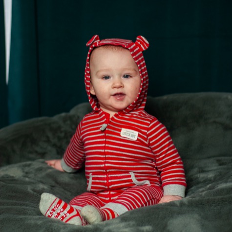 Professional baby portrait of a smiling infant wearing a red and white striped hooded outfit while seated on a soft gray blanket