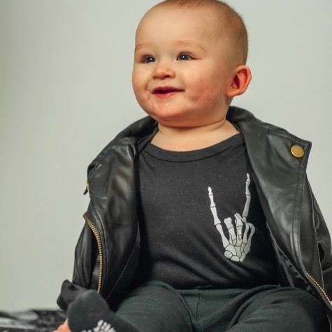 Professional studio baby portrait of a smiling infant wearing a black jacket over a dark graphic shirt against a clean light background