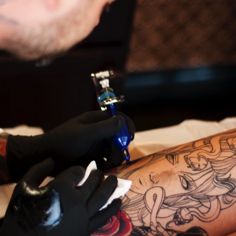Professional branding photo of tattoo artist Doren wearing black gloves and using a blue tattoo machine to work on a detailed illustrated arm tattoo