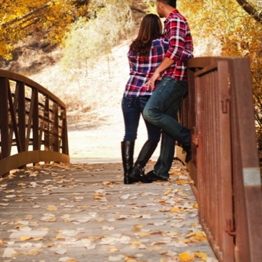A man and woman in matching red and blue plaid shirts and denim jeans embrace each other while standing on a wooden bridge during a couple's Valentine's Day portrait photography session by Kay Fritze of Fritze Media
