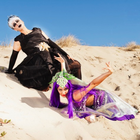 Two women in dramatic black and purple fantasy costumes performing expressive poses among sunlit coastal sand dunes during a creative professional portrait photography session by Kay Fritze of Fritze Media