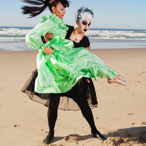Two women in theatrical green and black costumes performing a playful pose on a sandy ocean beach during a creative professional portrait photography session by Kay Fritze of Fritze Media