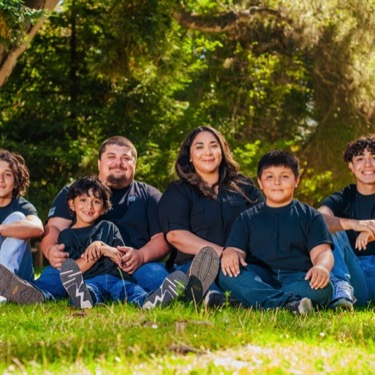 Family of six wearing coordinated black shirts and blue jeans seated together on grass at Miguelito Park during a professional family portrait photography session by Kay Fritze of Fritze Media