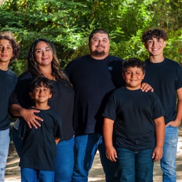 Family of six in coordinated black shirts and blue jeans standing together beneath trees at Miguelito Park during a professional family portrait photography session by Kay Fritze of Fritze Media