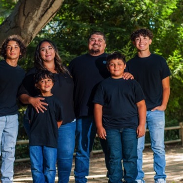 Parents and four sons wearing coordinated black shirts and blue jeans posing together outdoors at Miguelito Park during a professional family portrait photography session by Kay Fritze of Fritze Media