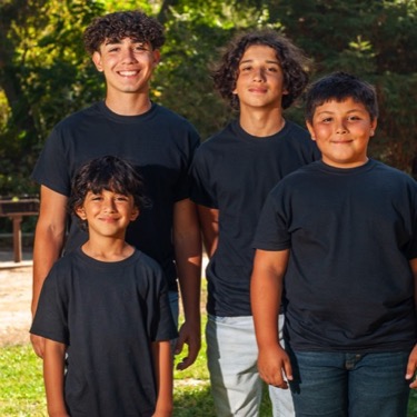 Four brothers wearing coordinated black shirts posing together on a sunlit lawn during an outdoor professional sibling portrait photography session by Kay Fritze of Fritze Media