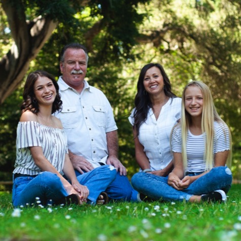 Family of four seated together on green grass beneath mature trees during an outdoor professional family portrait photography session by Kay Fritze of Fritze Media