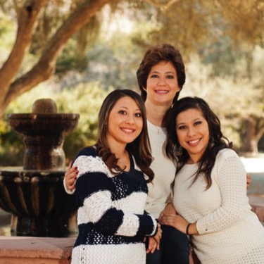 Three women from the same family standing closely together beside a stone fountain in a shaded outdoor setting during a professional family portrait photography session by Kay Fritze of Fritze Media