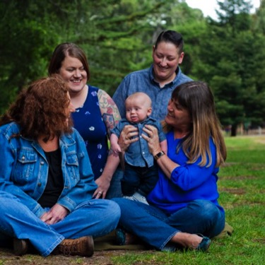 Four women gathered affectionately around a baby while seated on grass in a wooded park during a professional multigenerational family portrait photography session by Kay Fritze of Fritze Media