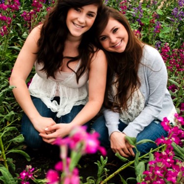 Two sisters seated closely together among bright pink flowers and greenery during an outdoor professional sibling portrait photography session by Kay Fritze of Fritze Media