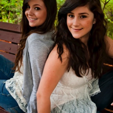 Two sisters smiling while seated together on a wooden park bench during an outdoor professional sibling portrait photography session by Kay Fritze of Fritze Media