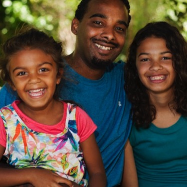 Father smiling with his two daughters in a close outdoor family portrait beneath leafy trees during a professional family portrait photography session by Kay Fritze of Fritze Media