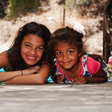 Two sisters lying side by side and smiling toward the camera outdoors during a playful professional family portrait photography session by Kay Fritze of Fritze Media