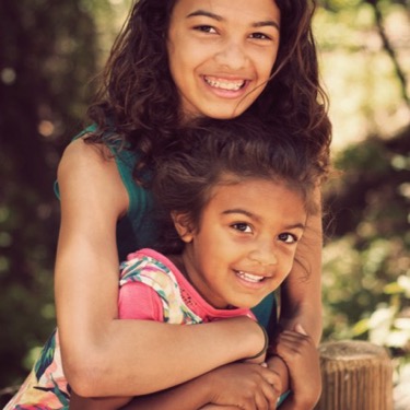Older sister hugging her younger sister from behind while both smile outdoors during a professional family and sibling portrait photography session by Kay Fritze of Fritze Media