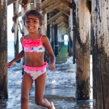 Young girl in a colorful swimsuit smiling and running in the sand beneath a rustic wooden pier during an outdoor professional child portrait photography session by Kay Fritze of Fritze Media