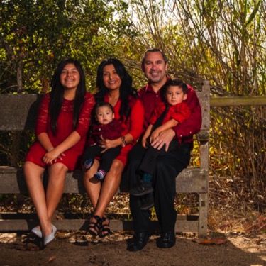 Family of five wearing coordinated red and black clothing seated together on a rustic wooden bench during an outdoor professional family portrait photography session by Kay Fritze of Fritze Media