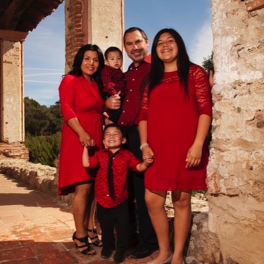 Family of five wearing coordinated red and black outfits standing together beside a rustic stone wall during an outdoor professional family portrait photography session by Kay Fritze of Fritze Media