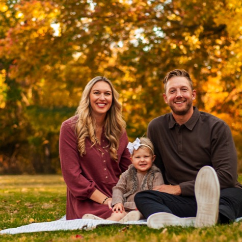 Mother and father seated with their young child beneath golden fall foliage during an outdoor professional family portrait photography session by Kay Fritze of Fritze Media