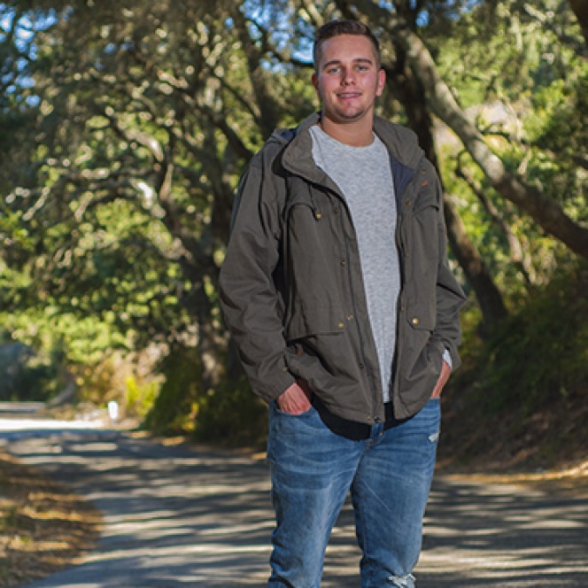 Young man in a dark jacket standing on a tree-lined path during an outdoor professional portrait photography session by Kay Fritze of Fritze Media