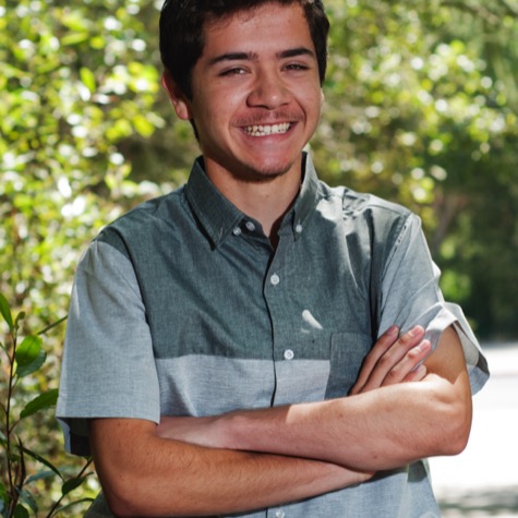 Professional outdoor portrait of a smiling young man in a gray button-up shirt standing with his arms crossed against a softly blurred green woodland background