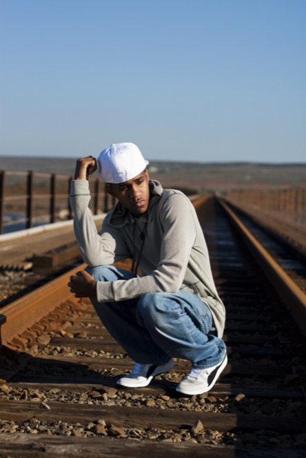 Young man in a gray hoodie and white cap crouching on railroad tracks during a dramatic outdoor portrait session by Kay Fritze of Fritze Media
