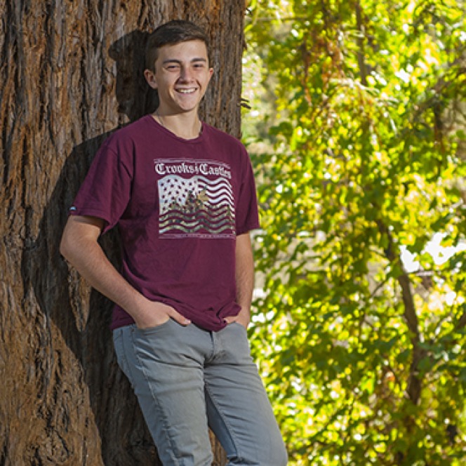 Young man in a maroon shirt posing beside a large tree during an outdoor professional senior portrait session by Kay Fritze of Fritze Media