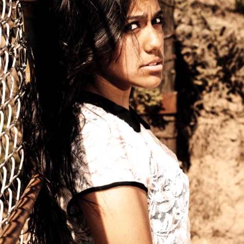 Dramatic outdoor portrait of a young woman with long dark hair posing beside a chain-link fence and weathered wall in strong sunlight