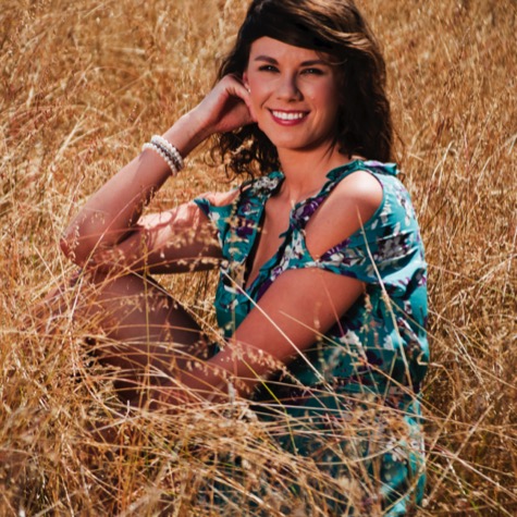 Professional outdoor portrait of a smiling woman in a patterned turquoise top seated among tall golden grass in a sunlit field