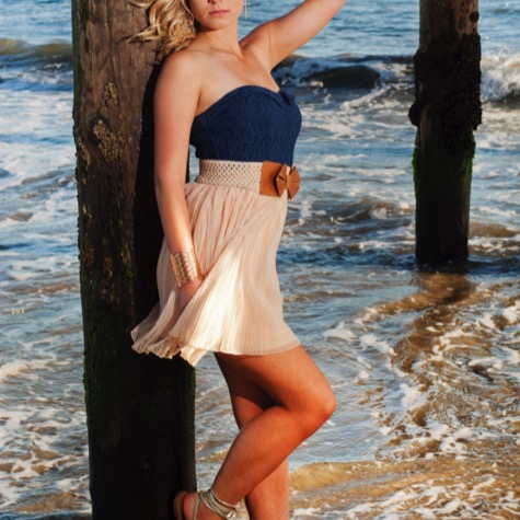 Professional fashion portrait of a blonde woman in a strapless blue and cream dress posing against a wooden pier post in shallow ocean surf