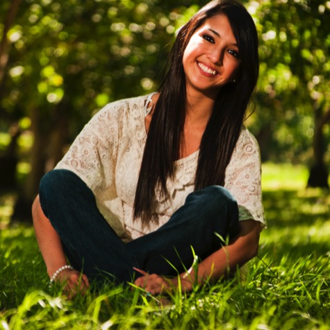 Professional outdoor portrait of a smiling woman with long dark hair seated cross-legged on green grass beneath sunlit trees
