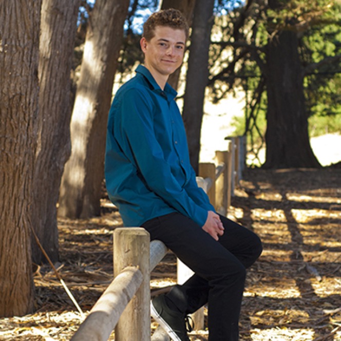 Young man in a blue shirt posing beside a wooden fence in a wooded setting during a professional senior portrait session by Kay Fritze of Fritze Media