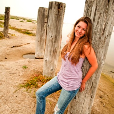 Young woman in a pink top and jeans posing beside rustic wooden posts during an outdoor professional portrait session by Kay Fritze of Fritze Media