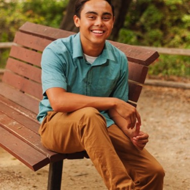 Young man in a teal polo and tan pants seated on a park bench for professional outdoor portrait photography by Kay Fritze of Fritze Media