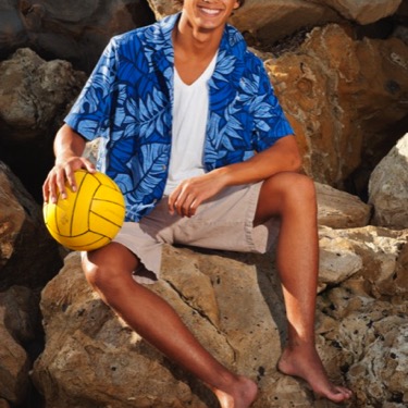 Young man in blue Hawaiian shirt holding a yellow volleyball on coastal rocks during a professional portrait photography session by Kay Fritze of Fritze Media